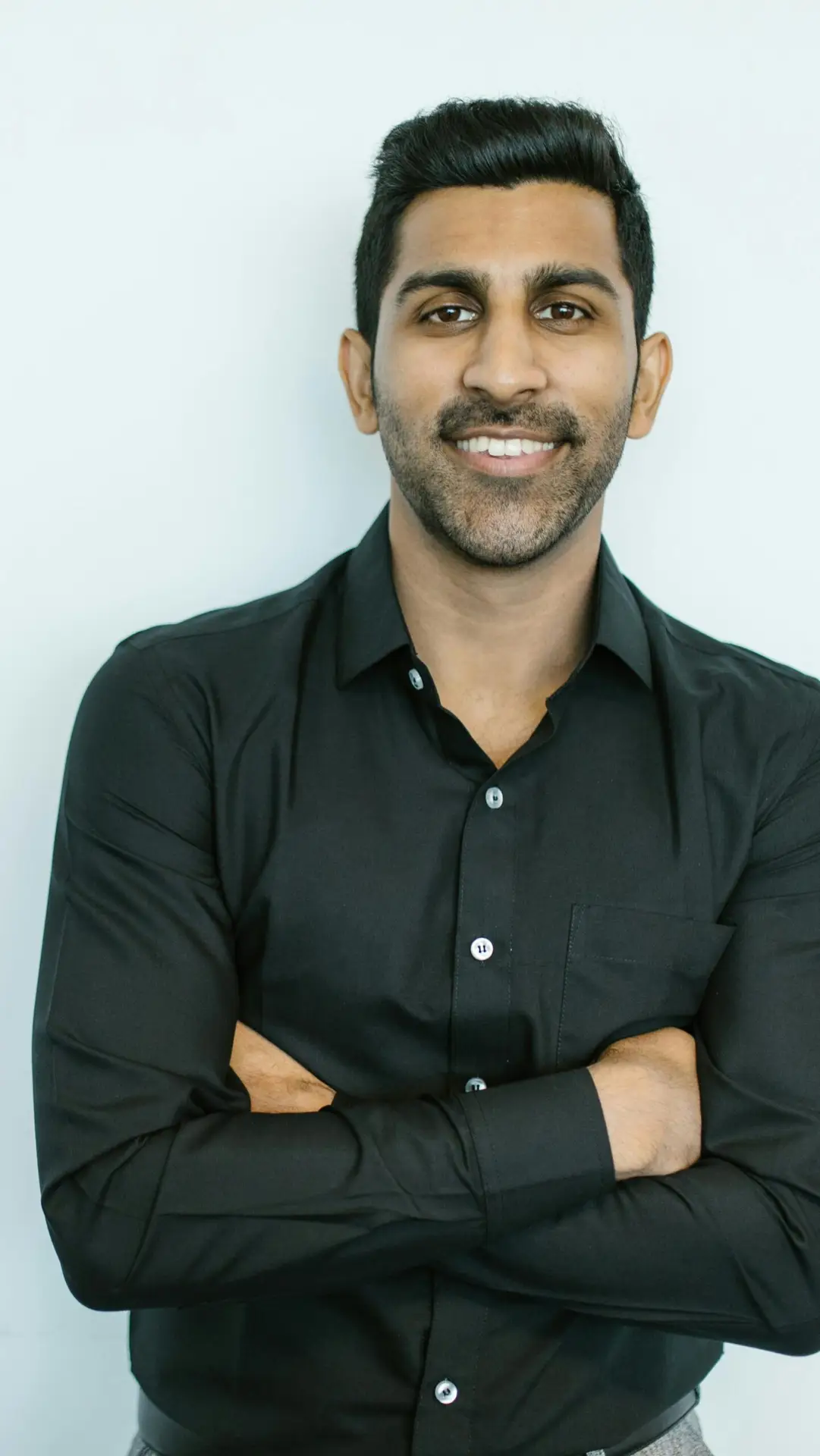 Portrait of a confident man in black shirt smiling against a plain background.